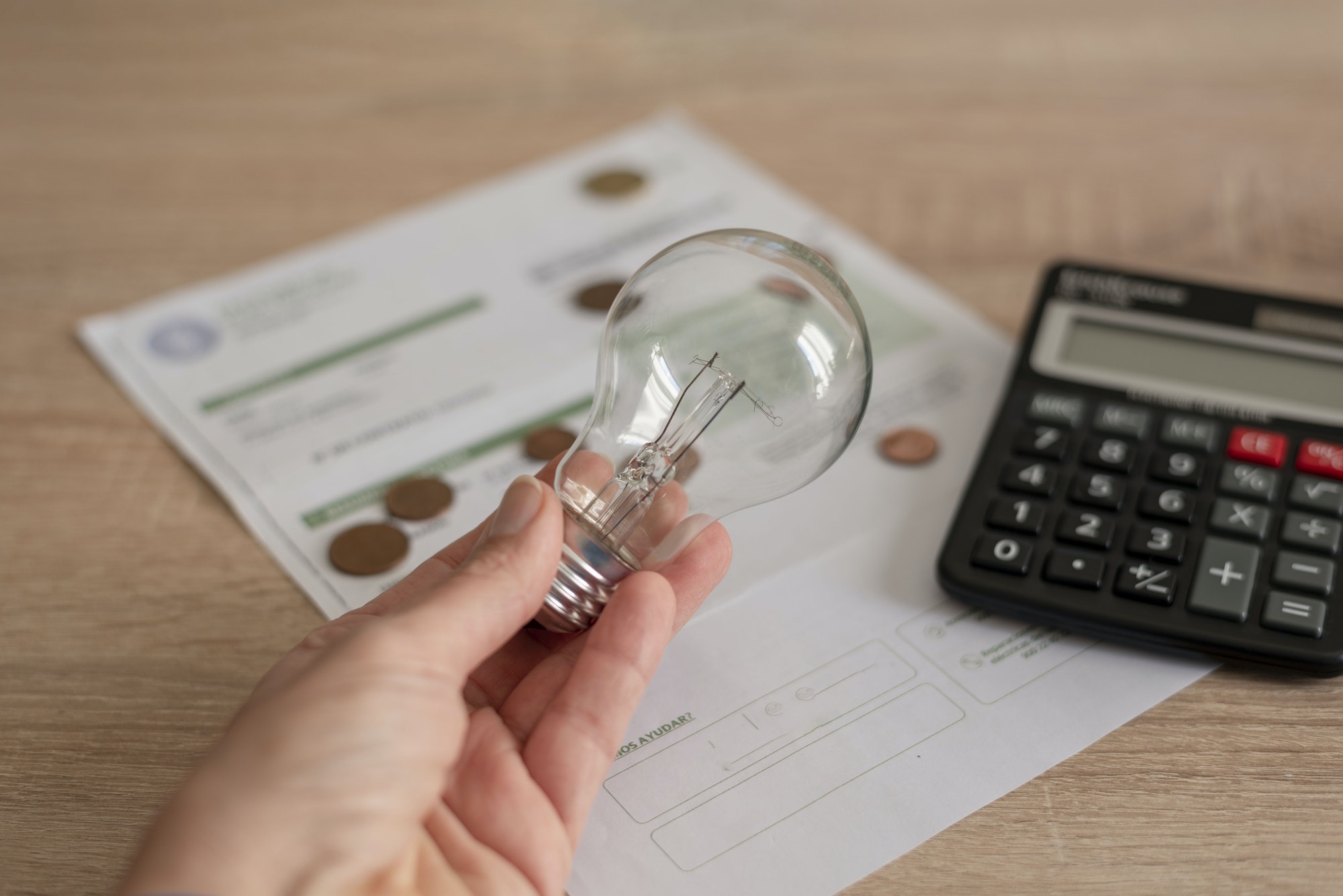 One hand holding light bulb over electricity bill, several coins and a calculator on the bill.